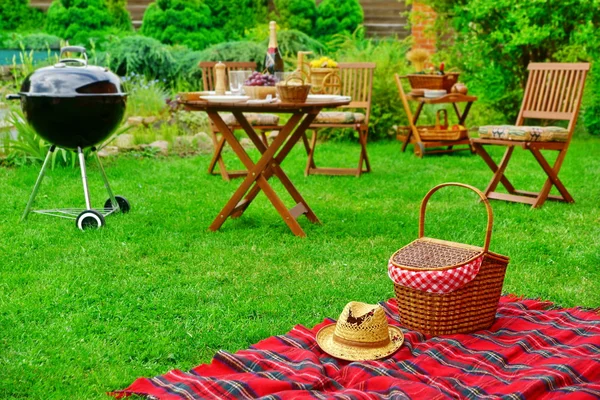 Closeup Of Red Picnic Blanket With Straw Hat And Basket Or Hamper. Blurred Outdoor Wooden Furniture In The Background. Family Home Backyard BBQ Party Or Picnic Conceptual Scene