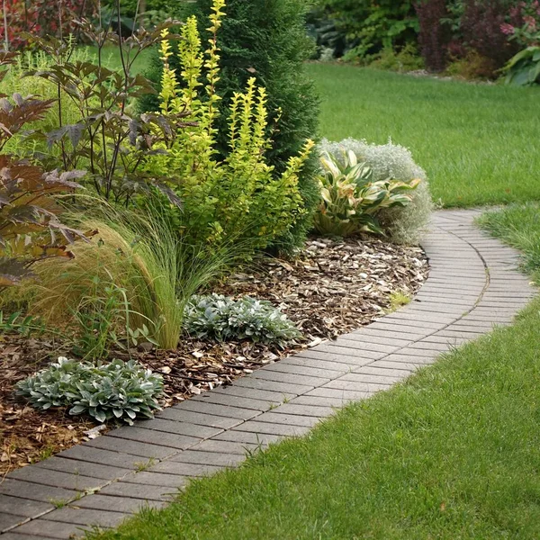 Backyard Garden Modern Design Landscaping. Decorative Garden Winding Pathway Walkway From Black Bricks. Back Yard Lawn And Natural Mulched Border Between Grass And Curved Brick Paving. Path To House.