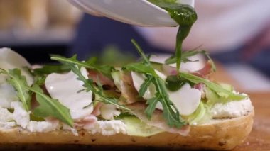 Ev mutfağında yemek hazırlıyorum. Kahvaltı hazırlamak ya da restoranda öğle yemeği. Fast food yakın plan. Erkek bir şef peynirli salatalık ve salatalı büyük bir sandviçin üzerine sos döker.