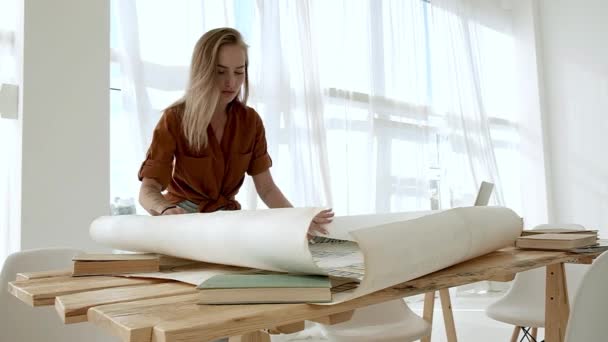 プロジェクトの巻物やドローイングでデスクで働く明るい広々としたオフィスの若い女の子が鉛筆を持ち 計画図を展開します 建築家 デザイナーの創作空間 — ストック動画