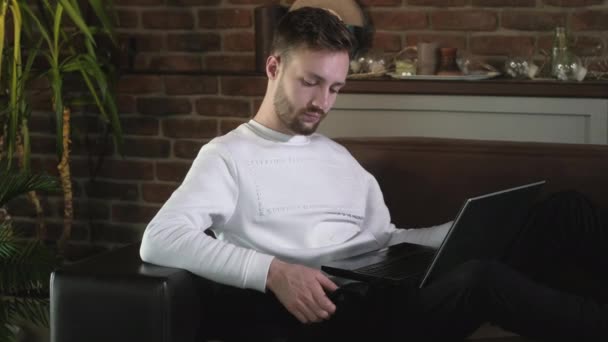 若い魅力的な男が黒い猫と居心地の良い家庭環境でソファに座っているとノートパソコンを入力しています ペットとの遠隔作業 隔離されたフリーランスは自宅で働いています — ストック動画