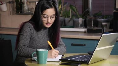 Evde, mutfakta Asyalı bir bayan öğrenci dizüstü bilgisayardan eğitim videoları izliyor. Kız bilgi aramak için internette geziniyor ve bir deftere yazıyor. Çevrimiçi uzaktan eğitim