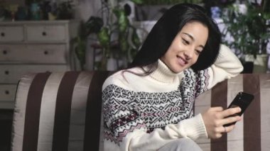 Oturma odasındaki kanepede süveter giyen genç Asyalı kadın akıllı telefon kullanıyor, sürpriz mesajlar okuyor, oturma odasındaki cep telefonundan iyi haberlerin tadını çıkarıyor. Mutlu kız telefonda iyi haberler okuyor.