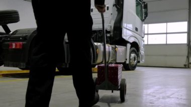 Siyah üniformalı oto tamircisi, atölyedeki kırmızı arabadaki beyaz bir kamyona iş aletleri ve yedek parça getiriyor. Otomobil servisi ve araba tamiri.