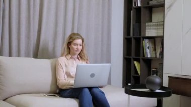 Sevimli genç kız parlak oturma odasında koltukta oturuyor ve modern dizüstü bilgisayar yazıyor. Genç serbest çalışan evde çalışır ve iletişim ve çevrimiçi olarak bilgisayar kullanır. İş kadını portresi ev ofisinde.