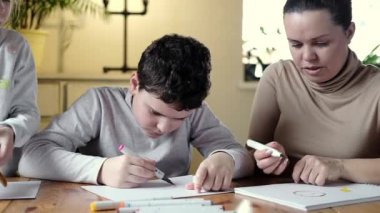 Yaratıcı çalışma odasında samimi ve mutlu bir aile. Genç anne ve kız ve oğlan kağıt masaya resim çiziyor. Anaokulu çocuk gelişimi kavramı. Çocuklara okul ve ev çizmeyi öğretmek