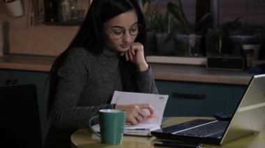 Genç ve çekici Asyalı kız öğrenci mutfakta oturmuş bilgisayarında ders çalışıyor. Kız bilgi aramak için internette geziniyor ve bir deftere yazıyor. Çevrimiçi uzaktan eğitim