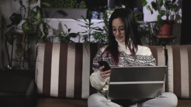 Genç Asyalı gözlüklü ve sıcak bir kazak giyen bir kadın akşamleyin dairenin oturma odasındaki kanepede oturuyor ve bir dizüstü bilgisayar üzerinde çalışıyor. Serbest çalışan bir kız evdeki bilgisayarda mesaj yazar.