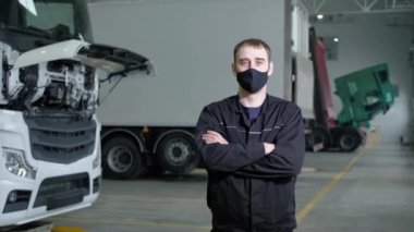 Tamir servisindeki genç profesyonel tamircinin portresi. Üniformalı ve koruyucu maskeli bir erkek mühendis bir tamirhanenin arka planında duruyor ve kameraya bakıyor..