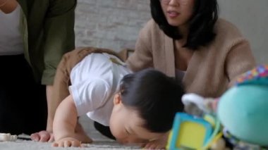 Asyalı bebek, annesi ve babasıyla odada yerde oturuyor. Çocuk ayağa kalkıp ilk adımı atmaya çalışır. Genç ebeveynler, oğullarının başarısı ve gelişimi için içtenlikle sevinirler.
