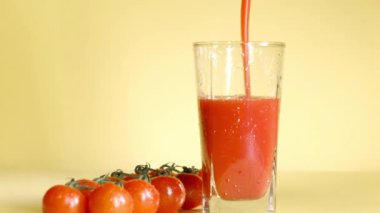 Domates suyu sarı arkaplandaki bir bardağa dökülür. Filiz üzerinde küçük domatesler yakınlarda uzanıyordu. Taze sebzeler sağlıklı yiyecek ve içecekler. Vitamin Kokteyli