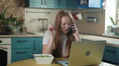 Mutfakta, evin içinde, modern bir telefonla konuşan, gülen ve bilgisayarla çalışan neşeli genç bir kadın. Dişi mutfakta bilgisayar kullanıyor ve sebze salatası yiyor.