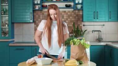 Evin mutfağında çekici genç bir kadın taze sebze salatalık salatası hazırlıyor. Olgun salatalık ve domates dilimleme işlemi. Masada salata malzemeleri var.