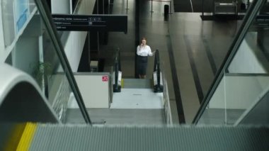 Yönetici genç iş kadını iş merkezindeki yürüyen merdivenin tepesine yürüyor. Bir bayan çalışan müşteriyle konuşmak için telefonu kullanır. en üstteki görünüm