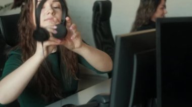 Merkezi ara. Kafkasyalı genç kadın müşteri destek uzmanı, bir kulaklık takıyor ve Çağrı Merkezi Kontrol Odası 'nda bir bilgisayar üzerinde çalışmaya başlıyor. Yardımcı operatör ofisteki bir bilgisayarda çalışıyor