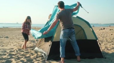 Kafkasyalı çocuk anne ve babasının kumlu deniz sahiline çadır kurmasına yardım ediyor. Tüm aile için açık hava aktiviteleri. Turist yaz sezonu, çadır ve sırt çantasıyla. piknik dışarıda