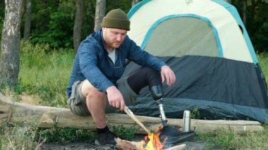 Kafkas engelli protez bacaklı adam ateşin yanındaki ormanda büyük bir kütüğün üzerinde oturuyor. İnsan çadır ve sırt çantasıyla seyahat eder. Kamp ateşinin yanında kamp yapmak. Turist ateşe odun atıyor.