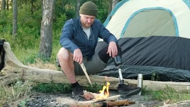 有假腿的白人残疾人正坐在靠近火堆的森林里的大木柴上 享受着孤独和休息 徒步旅行者带着帐篷和背包 在篝火边的大自然中露营 — 图库视频影像