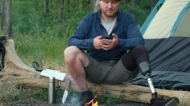 Ormanda protez bacaklı genç erkek turist uzun bir yürüyüşten sonra ateşin başında dinleniyor. Engelli kişi kütüğe oturur, alevden ısınır ve kameraya bakarak mesaj yazmak için telefonu kullanır