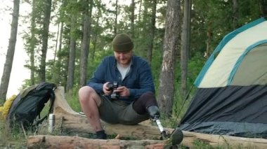 Protez bacaklı genç erkek turist uzun bir yürüyüşten sonra ormanda kamp ateşinde dinleniyor. Engelli kişi, güzel fotoğraflar yaratmak için kamera kullanarak kütüğün üzerinde oturuyor. Açık hava aktiviteleri