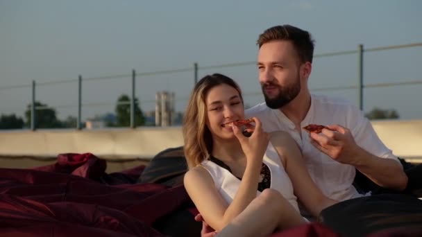 夏日黄昏的时候 年轻的白人男子和女孩正坐在柔软沙发上的屋顶上 人们吃披萨 玩得很开心 恋爱中的情侣在屋顶上约会 男朋友拥抱女朋友 — 图库视频影像