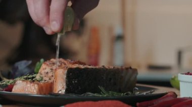 Sulu taze somon bifteği mutfak masasında sebzeler, otlar ve baharatlarla birlikte yatar. Şef bir dilim limonu deniz mahsulünün üzerine döküyor. Yemeklere lezzet katmak için..