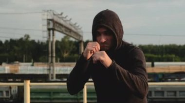 Kapüşonlu genç erkek sporcu saldırganca kameraya ve kutulara elleriyle bakıyor. Akşamüstü çatıda atlet eğitimi. Kardiyo egzersizleri. Dövüş için boksör trenleri