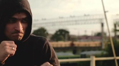 Kapüşonlu genç erkek sporcunun yakın çekimi kameraya ve kutulara elleriyle saldırarak bakıyor. Akşamüstü çatıda atlet eğitimi. Kardiyo egzersizleri. Dövüş için boksör trenleri