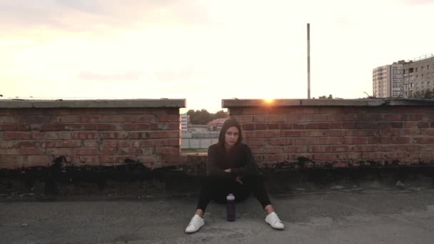 スポーツフーディの若い女性選手は 夏の日没を背景に訓練した後 レンガの壁の近くの屋根の上に座っている 女は屋根の上で運動した後に休んでいる — ストック動画