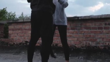 Kapüşonlu erkek boksör, gün batımının arka planında, şehir merkezinde egzersiz yapmak için kadının yumruğuna siyah bandaj sarar. Çatıda idman yapıyorum. Antrenör eğitimden önce kadına yardım eder