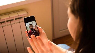 Genç esmer kadın akıllı telefonuyla görüntülü görüşme yapıyor, arkadaşıyla hoş sohbetler ediyor, evde dinlenirken ona selam veriyor. Sosyal uzaklık. Seçici odak
