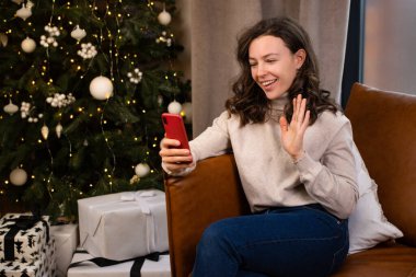 Genç ve mutlu esmer kadın akıllı telefonuyla video görüşmesi yapıyor. Arkadaşına ya da ebeveynine selam veriyor. Evdeki Noel ağacının yanındaki kanepede oturuyor. Çevrimiçi sohbet, yeni yıl kutlaması.