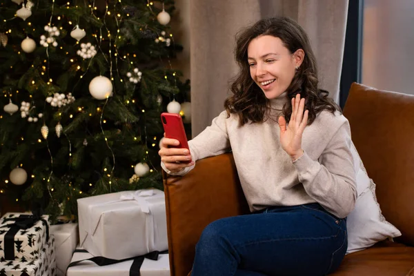 Genç ve mutlu esmer kadın akıllı telefonuyla video görüşmesi yapıyor. Arkadaşına ya da ebeveynine selam veriyor. Evdeki Noel ağacının yanındaki kanepede oturuyor. Çevrimiçi sohbet, yeni yıl kutlaması.