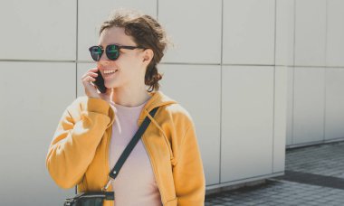 Gülümseyen hippi kadın cep telefonuyla konuşuyor sarı şık kapüşonlu ve güneş gözlüklü şehir merkezinde gri duvarın arkasında duruyor. Akıllı telefon kullanmak iyi sohbetti. Boşluğu kopyala