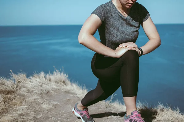 Sportif kadın bacaklarını esnetiyor ve kaslarını çalıştırıyor. Mavi deniz arka planında uçurumun kenarında koşmadan önce. Antrenmandan önce kas ve bağları hazırlıyorum. Boşluğu kopyala