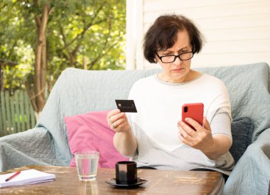 Kredi kartı tutan, cep telefonuna bakan, kanepe terasında oturan kafası karışmış son sınıf öğrencisi bir kadın. Borç sorunları, güvensiz çevrimiçi ödeme, başarısız işlem, para kaybı