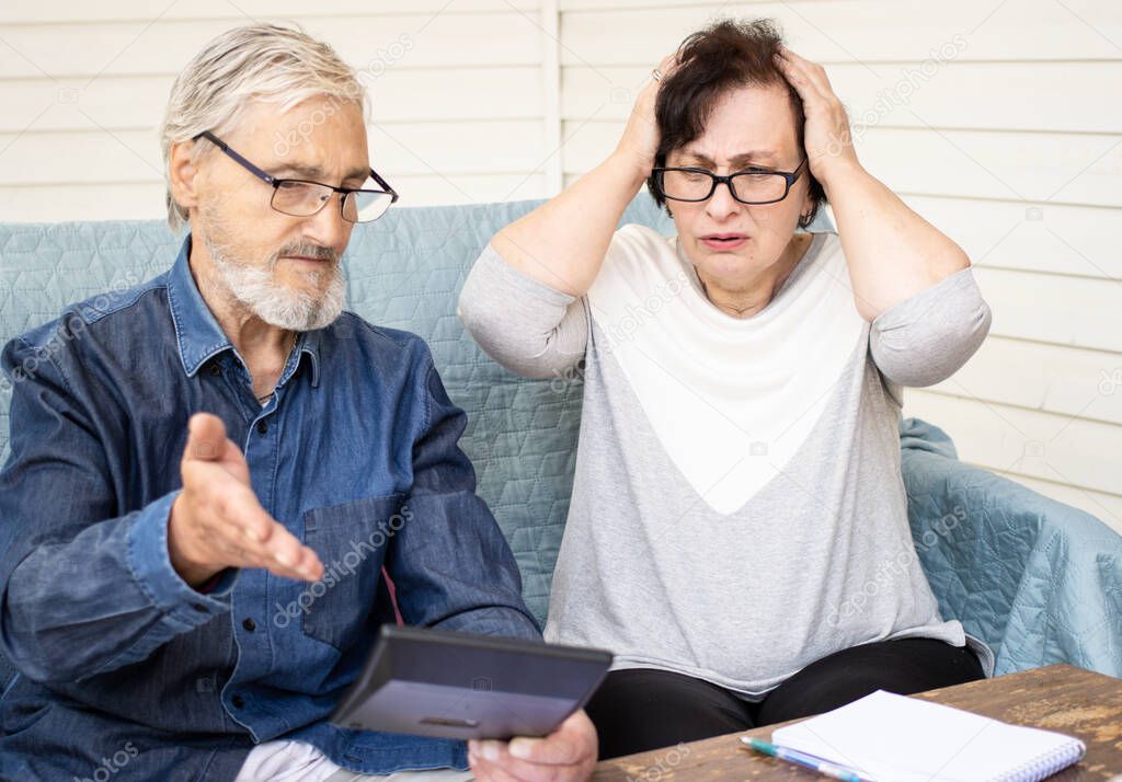 Little savings concept. Scared and worried mature couple looking at calculator, counting bank loan payment, calculating bills managing domestic finances. Worried about bankruptcy or money problem