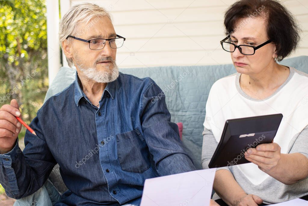 Scared shocked mature family couple looking at calculator counting loan payment, calculating bills and money from retirement, checking domestic finances, worried about bankruptcy or money problem