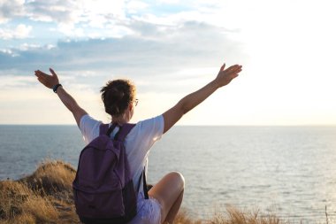 Sırt çantalı, dağ zirvesinde kollarını açmış, gün batımında kayanın üzerinde oturup denize bakan mutlu genç kadın yürüyüşçünün arka görüntüsü. Güçlü ve özgürlük yaşam tarzı konsepti. Macera gezisi.