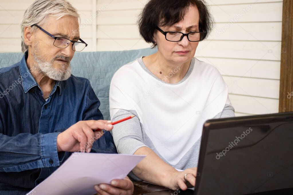 Focused elderly mature couple calculating bills and money from retirement using laptop, counting loan payment, checking domestic finances, sitting on home terrace. Pay utility bills via internet.