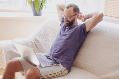 Gözlüklü mutlu bir iş adamı kollarını uzatıp, online işten sonra dizüstü bilgisayara bakıyor, evdeki kanepede oturuyor. Günün sonunun keyfini çıkarıyor, projenin başarıyla tamamlandığını hissediyor.