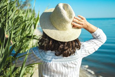 Yazlık hasır şapkalı esmer bir kadın ve kumsalda yeşil palmiye kamışlarından süzülen beyaz bir bez. Deniz kenarında ve gökyüzü arka planında. Seyahat, turizm, yeni rotalar keşfetmek, tatil kavramı.