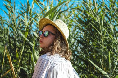 Yazlık hasır şapkalı kıvırcık esmer kadının kumsaldaki yeşil tropik palmiye ağaçlarının arasında dikilirken yan görüntüsü. Seyahat, yeni rotalar keşfetmek, yaz tatili 2021.