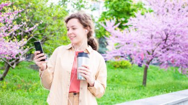Kulaklıkla gülümseyen genç bir kız öğrenci bahar aylarında açık hava parkında duran akıllı telefon ekranına bakıyor. Çevrimiçi webinar izlemek, dil öğrenmek, ses kurslarını dinlemek. Boşluğu kopyala