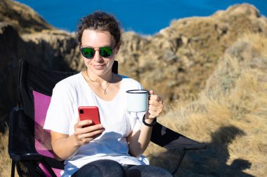 Akıllı telefon kullanan, elinde demir bardak kahve tutan, okyanus kenarındaki uçurumun kenarındaki turistik sandalyede oturan güzel bir kız gezgin. Sosyal medyayı kontrol eden, arkadaşlarıyla sohbet eden, haber okuyan, veri paylaşan.