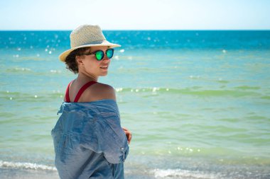 Yazlık hasır şapkalı ve kırmızı mayo giyen bir kadın kumsalda dikilip mavi deniz okyanusuna bakıyor. Yaz tatilinin tadını çıkarıyorum, tropik tatil beldesi. Uzayı kopyala Yaz tatili sezonu 2021