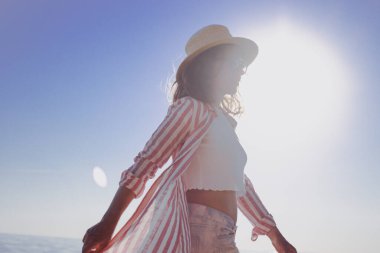 Hasır şapka ve açık renk kumaşla kadın siluetinin yan görüntüsü, deniz sahilinde yürümek, güneşli mavi gökyüzü arka planında yaz tatilinin tadını çıkarmak. Mutlu yaşam tarzı, seyahat macerası konsepti.