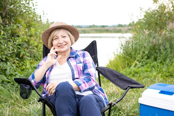 Beautiful elderly woman traveler in summer hat, smiling looking at camera, sitting in touristic chair near lake or river. Enjoying weekend picnic or camping vacation. Adventure lifestyle concept