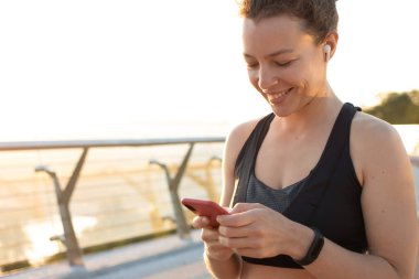 Akıllı telefon kullanarak kablosuz kulaklıkla koşan genç kadın antrenmandan sonra sosyal medyada çalışan verileri paylaşıyor, online fitness videosuna başlıyor. Kişisel antrenörle uzaktan eğitim. Boşluğu kopyala