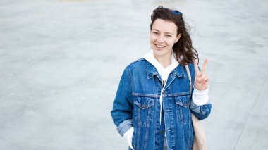 İyimser iyimser esmer kadın kameraya bakıp gülümsüyor barış jestleri yapıyor, zafer işareti yapıyor gri arka planda dikiliyor, hippi kot pantolon giyiyor. Boşluğu kopyala